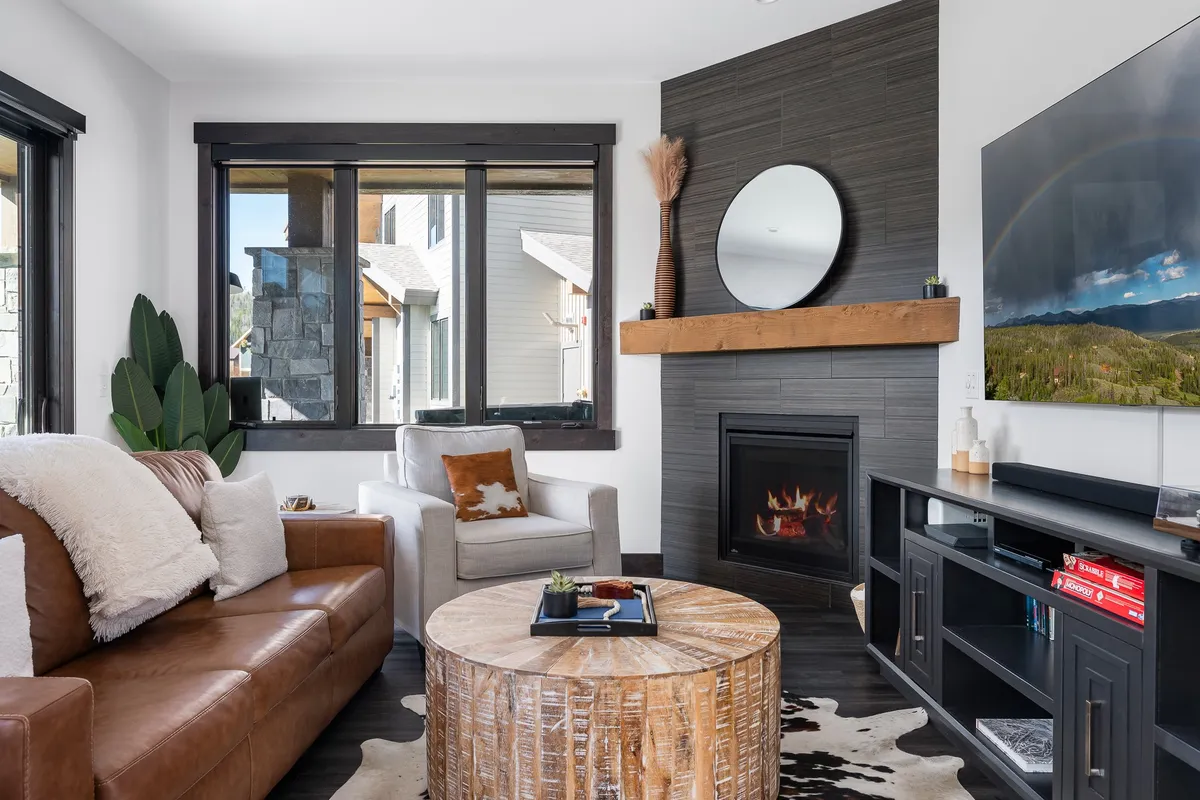 This image captures a spacious living room with a leather sofa, wooden coffee table, cozy fireplace, striking artwork, and large windows letting in natural light. The arrangement of plush seating and the warm glow from the fireplace create an inviting ambiance for relaxation or gathering with friends and family. The expansive windows also hint at scenic mountain views.