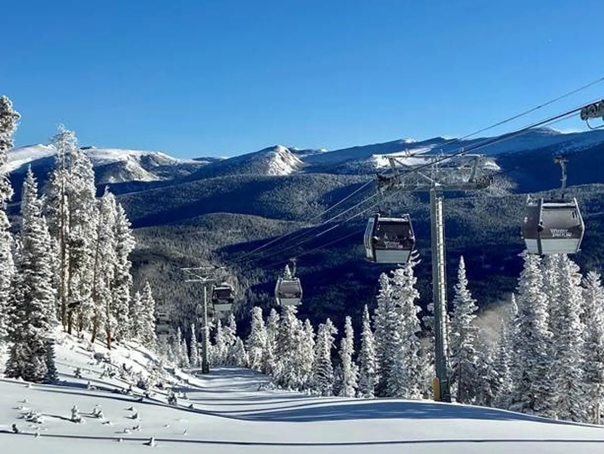An exterior view showcasing a ski lift ascending a snow-covered mountain, framed by tall pine trees and a dramatic mountain backdrop. The scene is lively, capturing the essence of Winter Park’s world-class skiing and outdoor adventure opportunities.