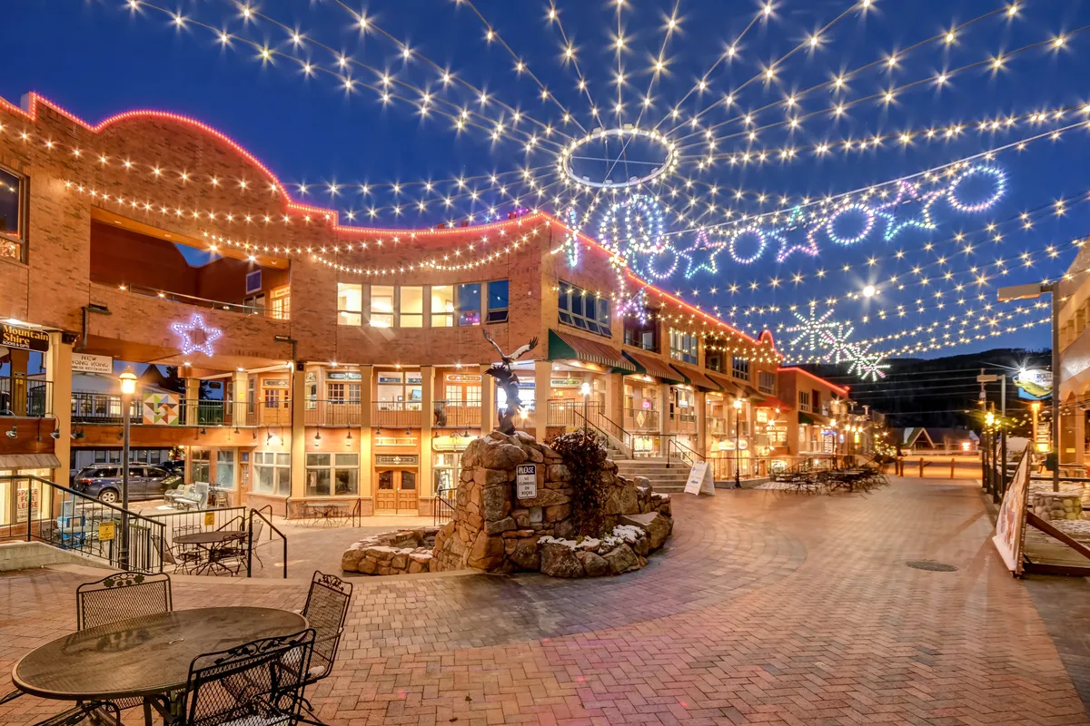 An enchanting evening exterior with twinkling lights strung overhead, decorative star shapes, and a stone fountain centerpiece. The inviting scene captures the festive, community-oriented atmosphere of downtown Winter Park, perfect for strolls, dining, or holiday shopping.