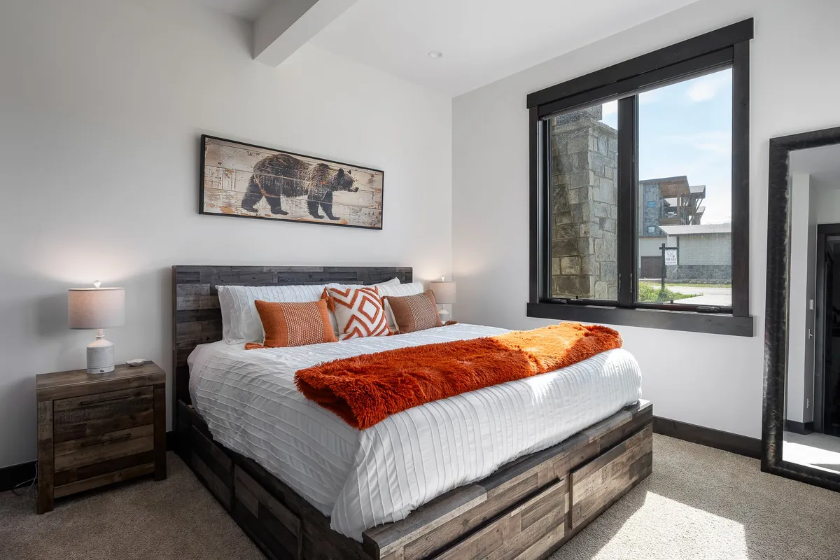 A cozy bedroom featuring a king bed with an orange throw blanket, bear-themed wall art, and a large window that lets in soft natural light. The mood exudes relaxation and comfort, emphasizing the room’s inviting atmosphere and restful vibe.
