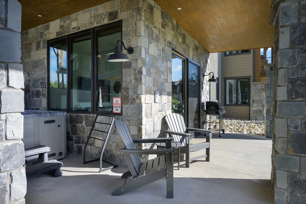 An inviting exterior setting showcases Adirondack chairs, a bubbling hot tub, stone walls, and a grill, all basking in the glow of sunset. This outdoor area is perfect for relaxing after a day of activities, gathering with family, or soaking under the stars.