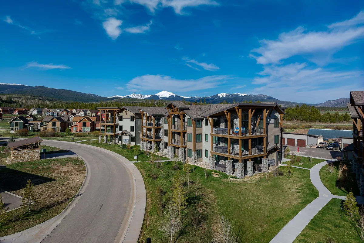 An inviting exterior view of the property with a stunning mountain range in the background, modern architectural lines, lush green landscaping, and a gently curving road leading towards outdoor adventure. The scene is tranquil yet hints at easy access to hiking, biking, and exploration.