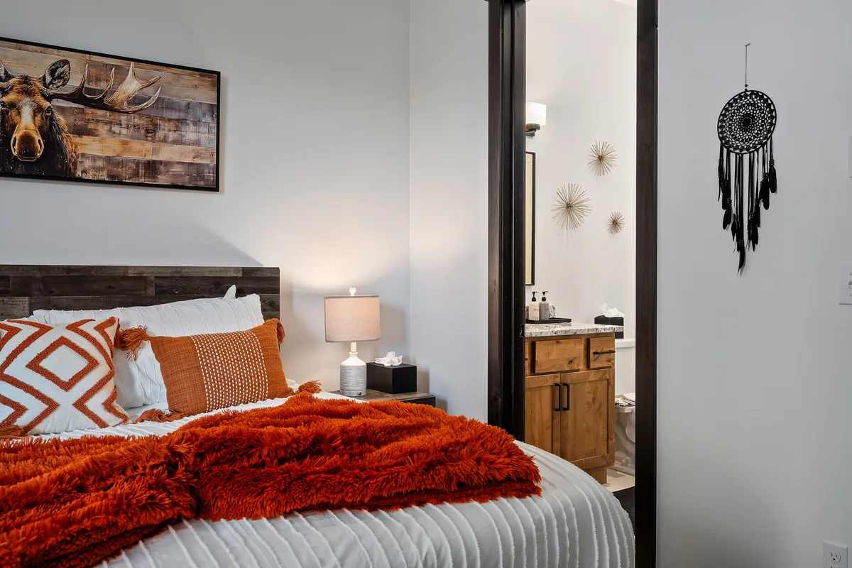 A beautifully appointed bedroom with a king bed accented by an orange throw blanket, tasteful moose artwork above the headboard, a dreamcatcher on the wall, and a glimpse of an ensuite bathroom. The space is brightened by large windows that provide abundant natural light, and the mood is welcoming and tranquil.